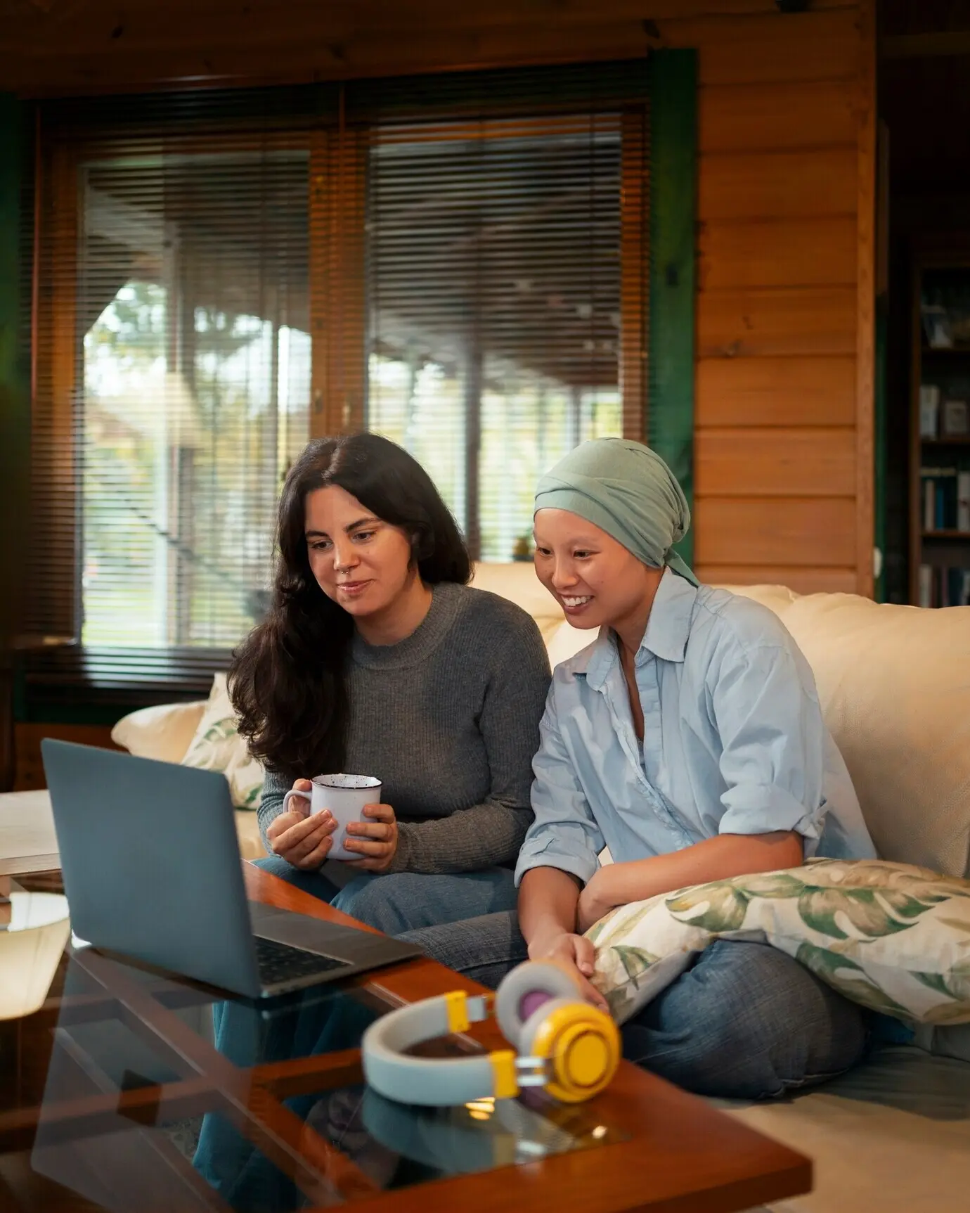 Porträt einer an Krebs erkrankten Frau, die zu Hause auf der Couch gemeinsam mit einer befreundeten Person einen Laptop benutzt.